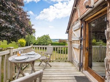 An outdoor wooden balcony with a round table set for two and wooden chairs overlooking a garden at The Cider Mill in Westhope near Leominster