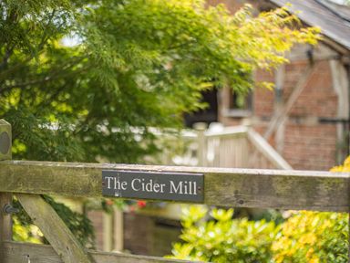 A wooden gate with a sign saying The Cider Mill and a tree and brick building in the background at The Cider Mill in Westhope near Leominster