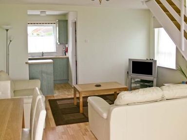 A living room with two cream sofas a wooden coffee table a TV on a stand and a kitchen with green cabinets visible at Seascape in Kessingland near Lowestoft