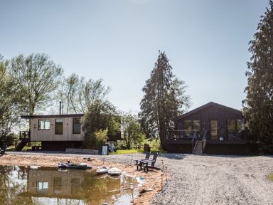 Two houses by the water with trees and gravel area at The Larch House in Farndon