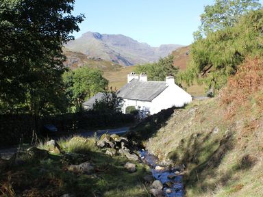 A cottage by a stream with mountains in the background at Owls Nook Lodge Langdale 9, White Cross Bay near Windermere