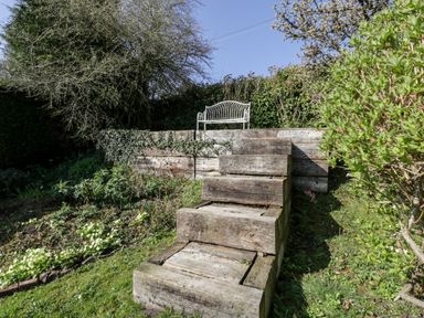 An outdoor garden area with wooden steps leading up to a white metal bench surrounded by plants and trees at Gordons View Pleasant Stile near Gloucester