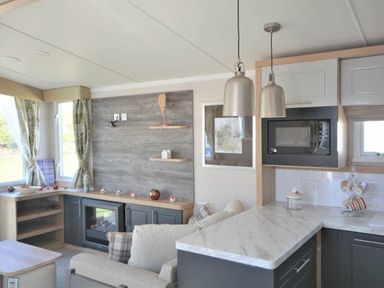 a living room with a grey sofa, a TV unit, a kitchenette with marble counter, and decorative shelves at Perran Sands - Holiday Accommodation 15554 in Perranporth