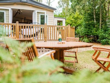 a wooden table and chairs with a view of a cabin and trees at Kelling Heath Holiday Park - Holiday Accommodation 18664 in Holt