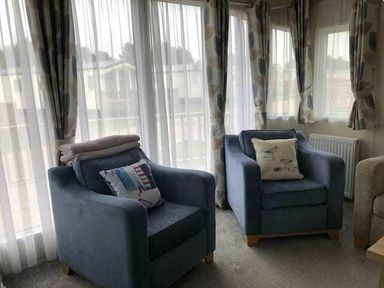 two blue armchairs with cushions and a stack of blankets in a room with large windows at Pinewoods Holiday Park - Holiday Accommodation 19734 Wells-Next-The-Sea