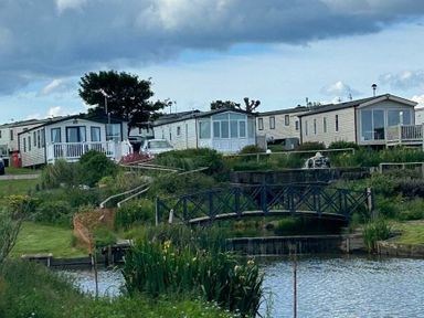 a group of holiday homes by a pond with a small wooden bridge at Sand Le Mere Holiday Village - Holiday Accommodation 20326 Tunstall