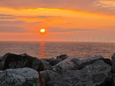 a sunset over the ocean with rocks in the foreground and wind turbines in the distance at Ty Gwyn Caravan Park - Holiday Accommodation 22189 Abergele