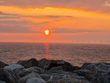a sunset over the ocean with rocks in the foreground and wind turbines in the distance at Ty Gwyn Caravan Park - Holiday Accommodation 22189 Abergele