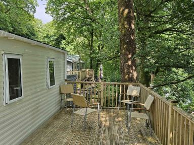 a deck area with chairs next to a mobile home in a wooded setting at Aberdunant Hall Holiday Park - Holiday Accommodation 22358, Porthmadog