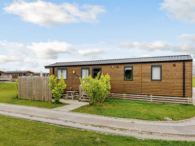 A wooden cabin with a fence and bushes in an outdoor area at Alder Waterside Spa in Carlton Miniott near Thirsk
