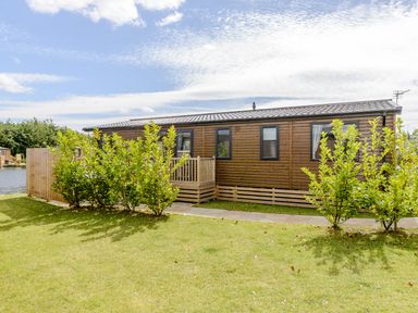 A wooden cabin with a deck and bushes at Oak Waterside Spa Carlton Miniott near Thirsk