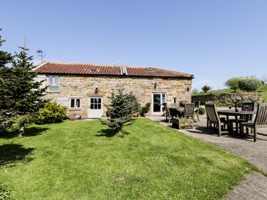 A stone house with a tiled roof a green lawn small trees and outdoor wooden dining tables at Honey Bee Cottage in Staintondale