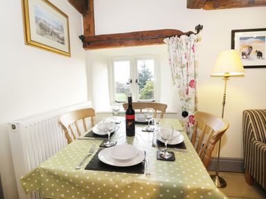 A dining table set with plates glasses and a bottle of wine near a window with floral curtains and a floor lamp in Honey Bee Cottage in Staintondale