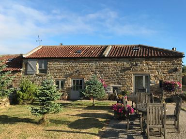 An outdoor seating area with wooden tables and chairs in front of a stone cottage with red tiled roof and small trees in the yard at Honey Bee Cottage in Staintondale