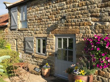 A stone cottage exterior with grey windows and door a small metal table and chairs and potted flowers at Honey Bee Cottage in Staintondale