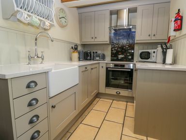 A kitchen with beige cabinets white countertops a white farmhouse sink a stainless steel oven and hood a dish rack with mugs toaster coffee maker microwave and fire extinguisher at Honey Bee Cottage in Staintondale