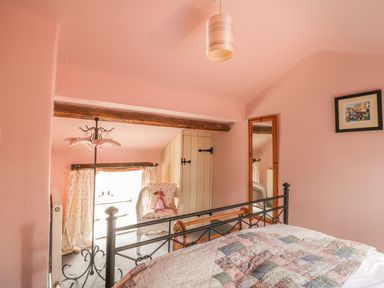 A bedroom with a metal bed frame a patchwork quilt a white wicker chair with a cushion and a doll a full-length mirror a small cupboard and a window with curtains at Honey Bee Cottage in Staintondale