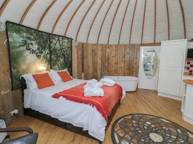 A bedroom with a double bed with orange pillows and blanket a bathtub a circular patterned rug and wooden walls at Lakeview Yurt Beckford near Tewkesbury