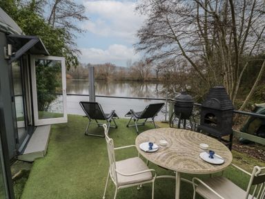 An outdoor patio with a mosaic table set, two reclining chairs, a barbecue grill, and a view of a lake surrounded by trees at Lakeview Yurt in Beckford near Tewkesbury
