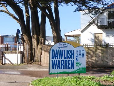 A sign for Dawlish Warren near trees and a building in Dawlish Warren