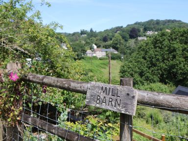 A sign on a fence indicating 'MILL BARN' with hills and trees in the background at The Mill Barn in Lustleigh
