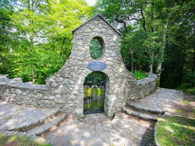 A stone archway with a metal gate and plaque in a forested area at Bron Eifion Pod 1 Criccieth