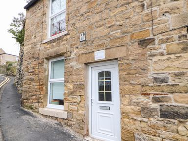 A stone exterior of a cottage with a white door and window next to a narrow road at Bridge Cottage in Haltwhistle near Hadrians Wall