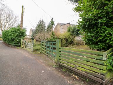 A road with green wooden fencing and gates leading to a garden with bushes and trees at Bridge Cottage in Haltwhistle near Hadrians Wall