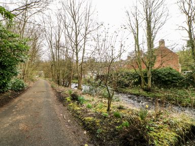 A pathway next to a river with trees and shrubs and houses in the background at Bridge Cottage in Haltwhistle near Hadrians Wall
