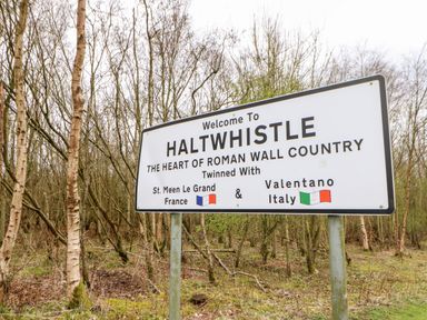 A welcome sign for Haltwhistle in a wooded area with trees and moss on the ground at Bridge Cottage in Haltwhistle near Hadrian's Wall