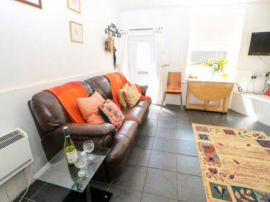A living room with a brown leather sofa with cushions and a red throw a glass table with a wine bottle and two glasses a wooden chair a foldable wooden table a wall-mounted television and a patterned rug at Bridge Cottage in Haltwhistle near Hadrian's Wall