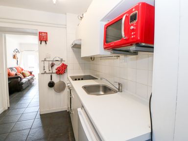 A narrow kitchen with a white countertop a red microwave a sink and utensils hanging on the wall at Bridge Cottage in Haltwhistle near Hadrian's Wall