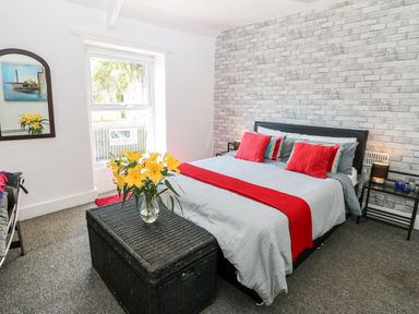 A bedroom with a bed with red and striped pillows a gray bedspread a black wicker chest at the foot of the bed with a vase of yellow flowers a window and a mirror on white walls at Bridge Cottage Haltwhistle near Hadrians Wall