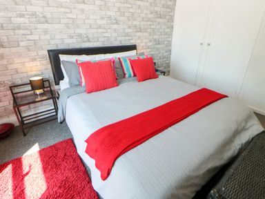 A bedroom with a bed with red pillows and a red throw on a grey cover next to a bedside table with a lamp at Bridge Cottage in Haltwhistle near Hadrian's Wall