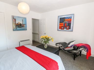 A bedroom with a bed featuring a red blanket a black chaise lounge with cushions and a red throw a table with yellow flowers two paintings on white walls at Bridge Cottage Haltwhistle near Hadrians Wall