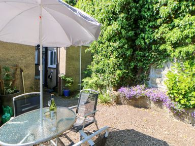 An outdoor patio area with a glass table and chairs under a large umbrella surrounded by plants and flowers at Bridge Cottage in Haltwhistle near Hadrian's Wall