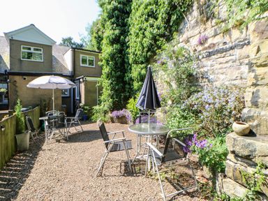 A garden patio with chairs tables umbrellas and plants by a stone wall at Bridge Cottage in Haltwhistle near Hadrians Wall
