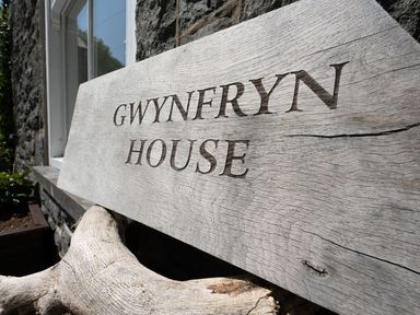 A wooden sign displaying Gwynfryn House outside a stone wall in Llanbedr
