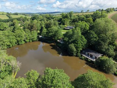 An aerial view of a pond surrounded by trees and buildings at 5 Tree Tops in Dobwalls