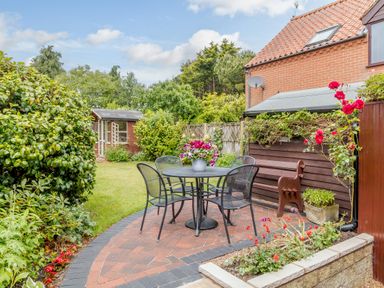 A garden with a round table and four chairs on a brick patio near a shed at Hollyhedge Cottage in Briston