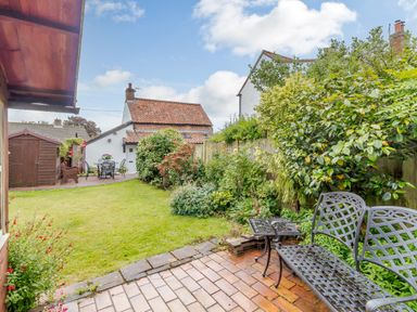 A garden with a metal bench and table on a paved area and a lawn with shrubs and plants at Hollyhedge Cottage in Briston
