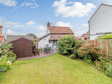 A garden with grass a wooden shed patio furniture and a small white house in the background at Hollyhedge Cottage in Briston