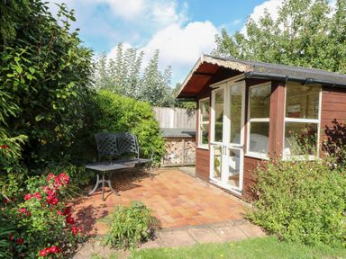 A garden shed with a paved patio and a bench at Hollyhedge Cottage in Briston