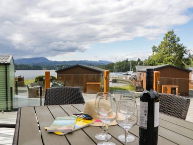 An outdoor area with a table, glasses, and a view of water and cabins at 19 Pony Meadow White Cross Bay near Troutbeck Bridge