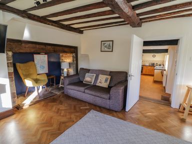 A living room with a gray sofa, a yellow chair, a side table with a lamp, wooden beams on the ceiling, and a kitchen visible through an open door at Little Cowarne Court in Little Cowarne near Bromyard