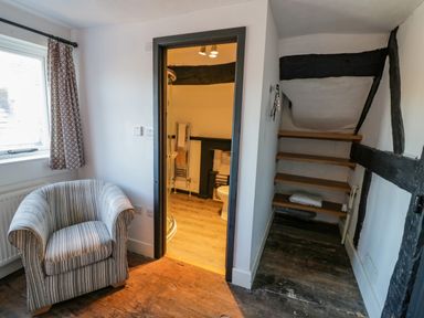 A room with a striped armchair near a window next to a doorway leading to a bathroom with a shower and toilet and wooden shelves in a corner at Little Cowarne Court in Little Cowarne near Bromyard