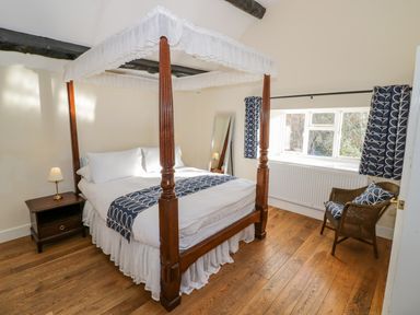A bedroom with a four-poster bed a bedside table with a lamp a wicker chair with cushions and a window with curtains at Little Cowarne Court in Little Cowarne near Bromyard