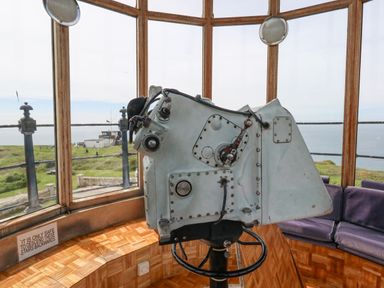 A light grey optical device mounted on a black stand inside a room with large windows showing a grassy coastal view at Old Higher Lighthouse Stopes Cottage Portland Bill