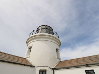 The top section of a white lighthouse tower with windows and railing under a partly cloudy sky at Old Higher Lighthouse Stopes Cottage in Portland Bill