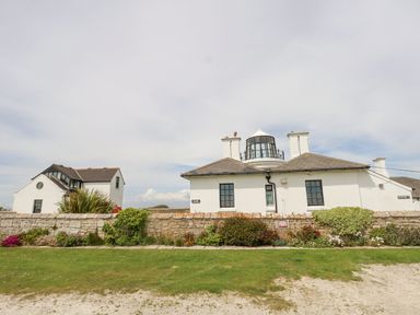 A white building with a lighthouse lantern room on top behind a stone wall with plants and grass at Old Higher Lighthouse Stopes Cottage in Portland Bill